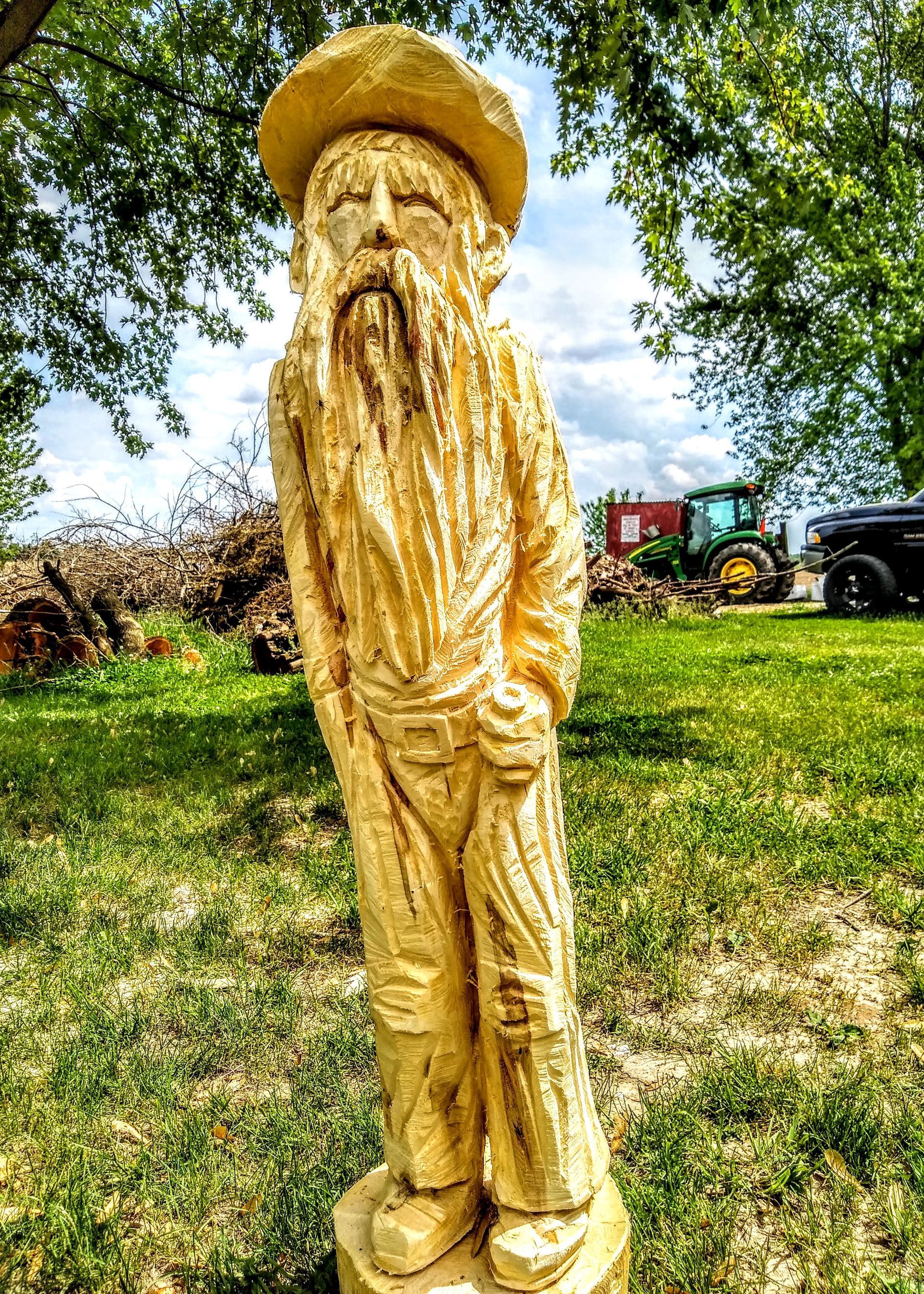 Wooden carved standing figure in a grassy field with trees and farm equipment in the background
