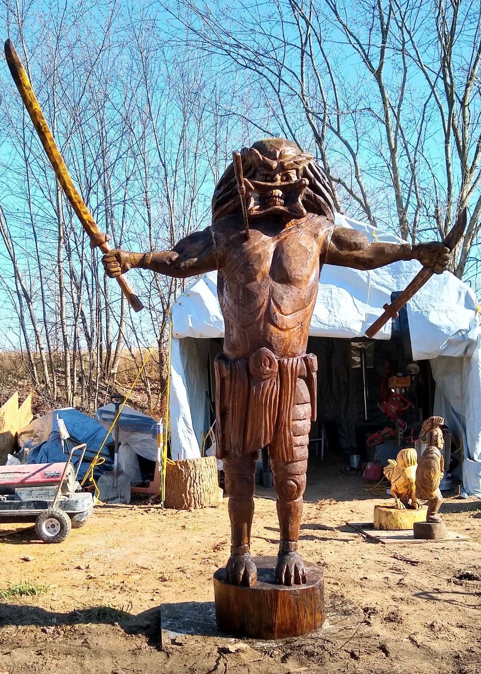 Wooden statue of a bearded figure with outstretched arms outdoors in a rustic yard