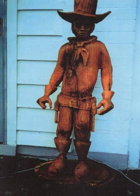 Wooden cowboy statue in a hat and boots standing against a light blue wall