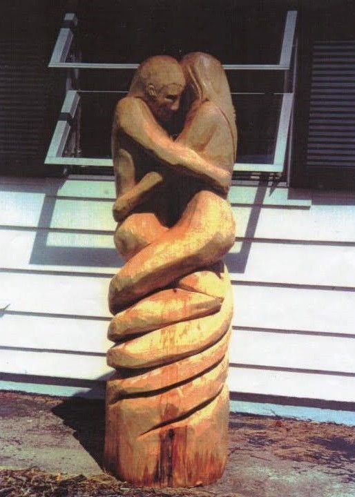Wooden sculpture of two figures embracing, with intertwined forms, standing outdoors against a building.
