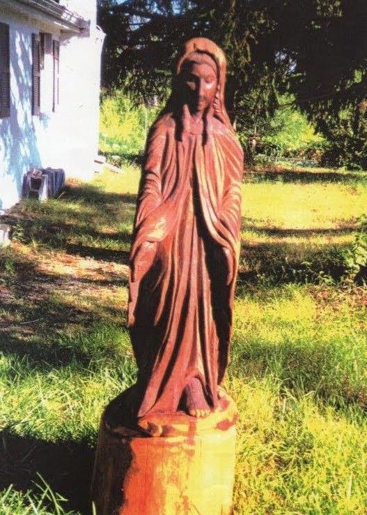 Weathered statue of a robed figure standing on a pedestal in a grassy yard beside a building.