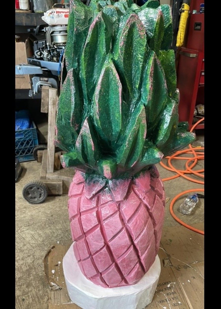 Pink and green pineapple sculpture standing in a workshop or garage, with a textured finish