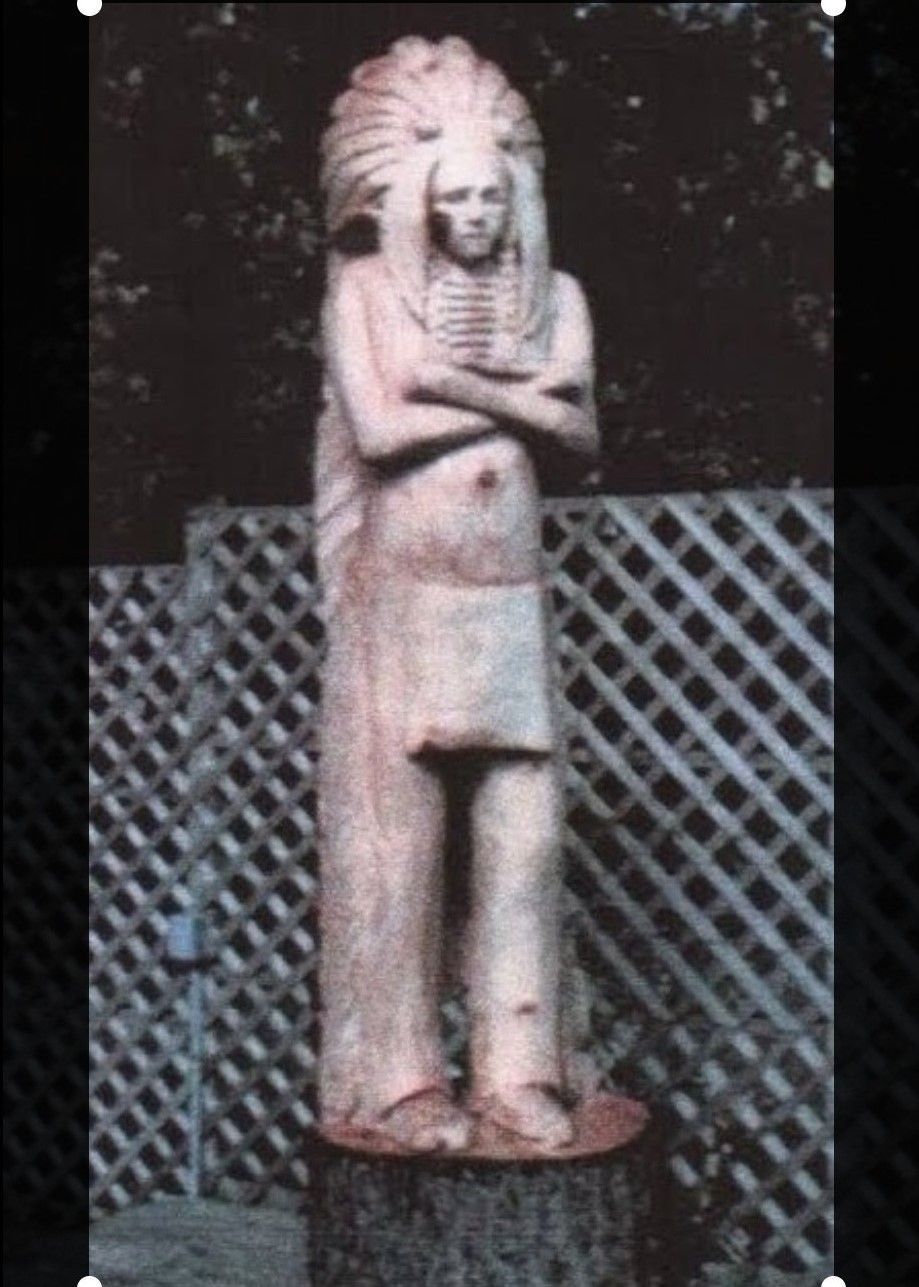 Stone statue of a standing figure with crossed arms in front of a lattice fence.