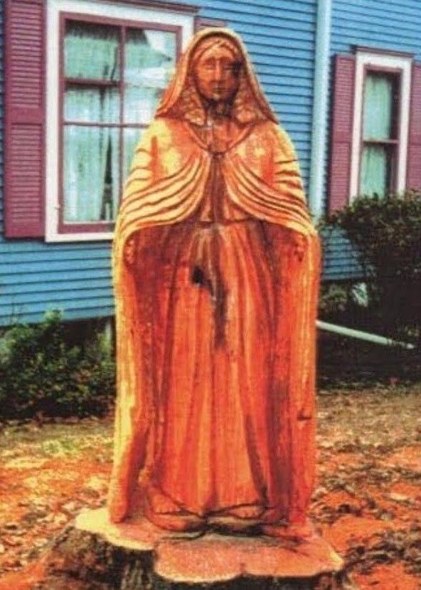 Wooden statue of a robed figure painted orange, standing in front of a blue house