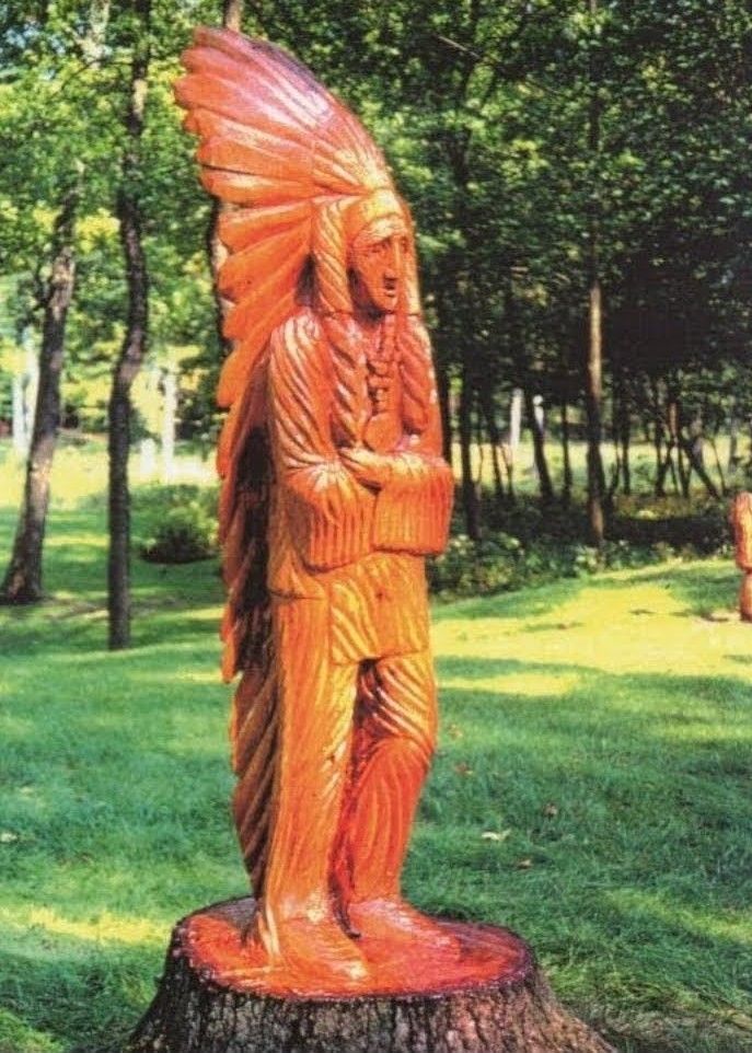 Orange wooden statue of a feathered figure standing in a grassy park among trees