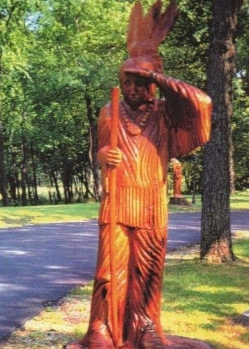 Wooden statue of a person in a feathered headdress standing beside a road in a wooded park