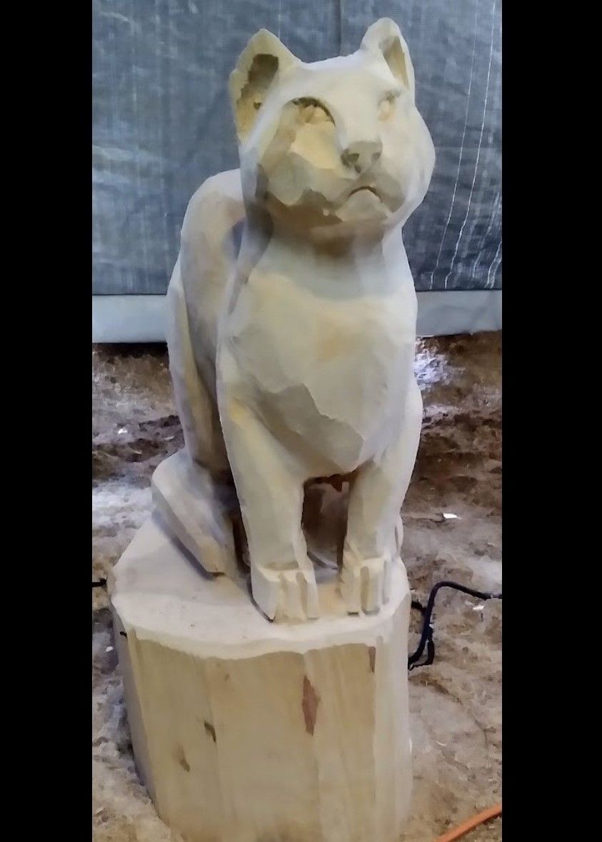 Carved wooden cat figurine on a stump base outdoors, with a pale unfinished surface.