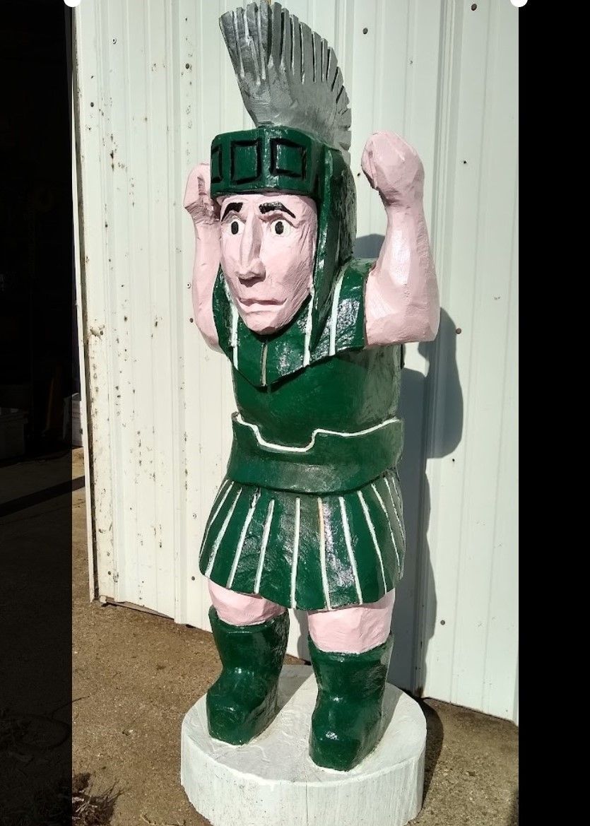 Green statue of a cheerleader or mascot with raised arms, standing on a white base outdoors.