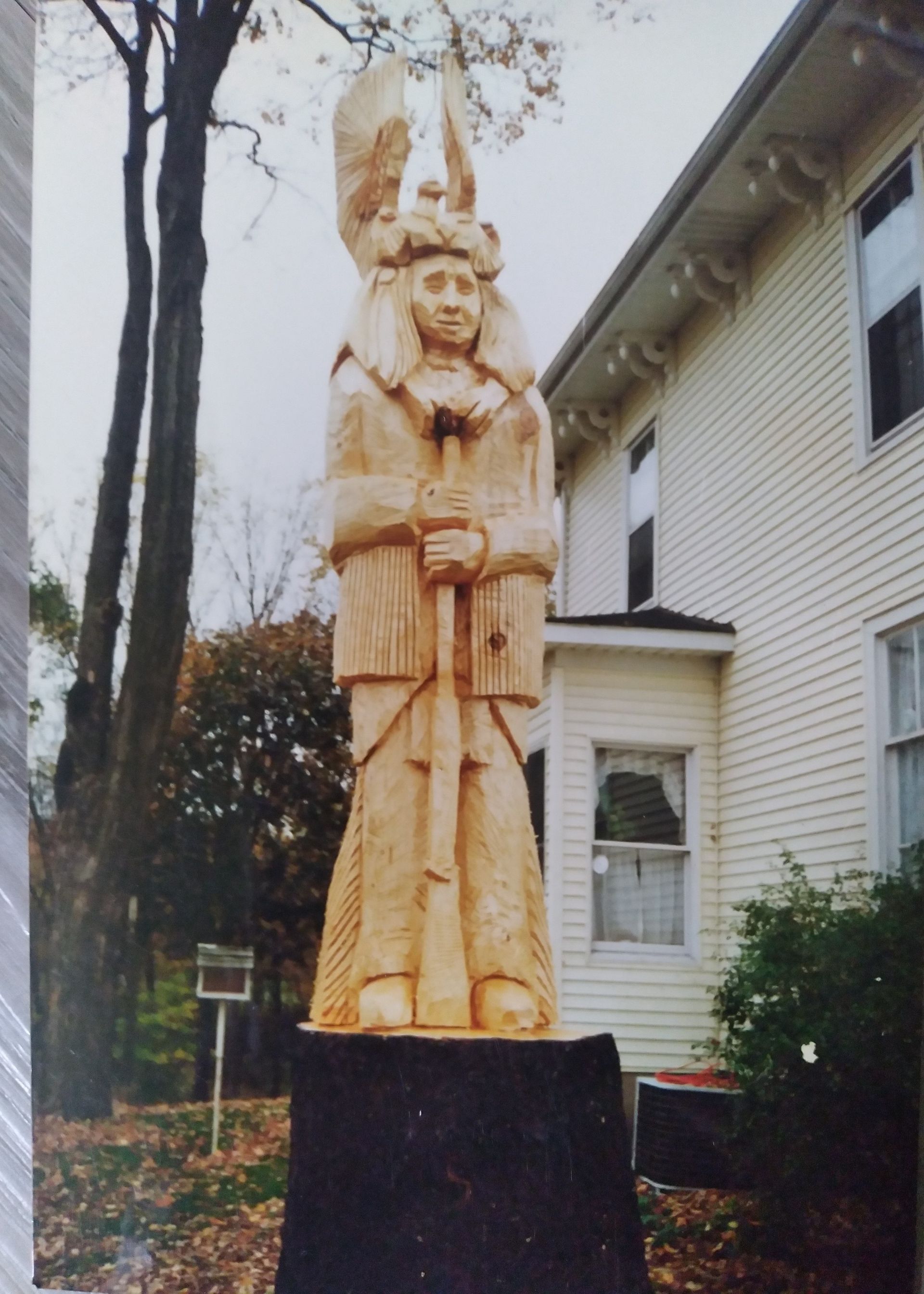 Wooden carved statue on a black pedestal beside a white house and trees