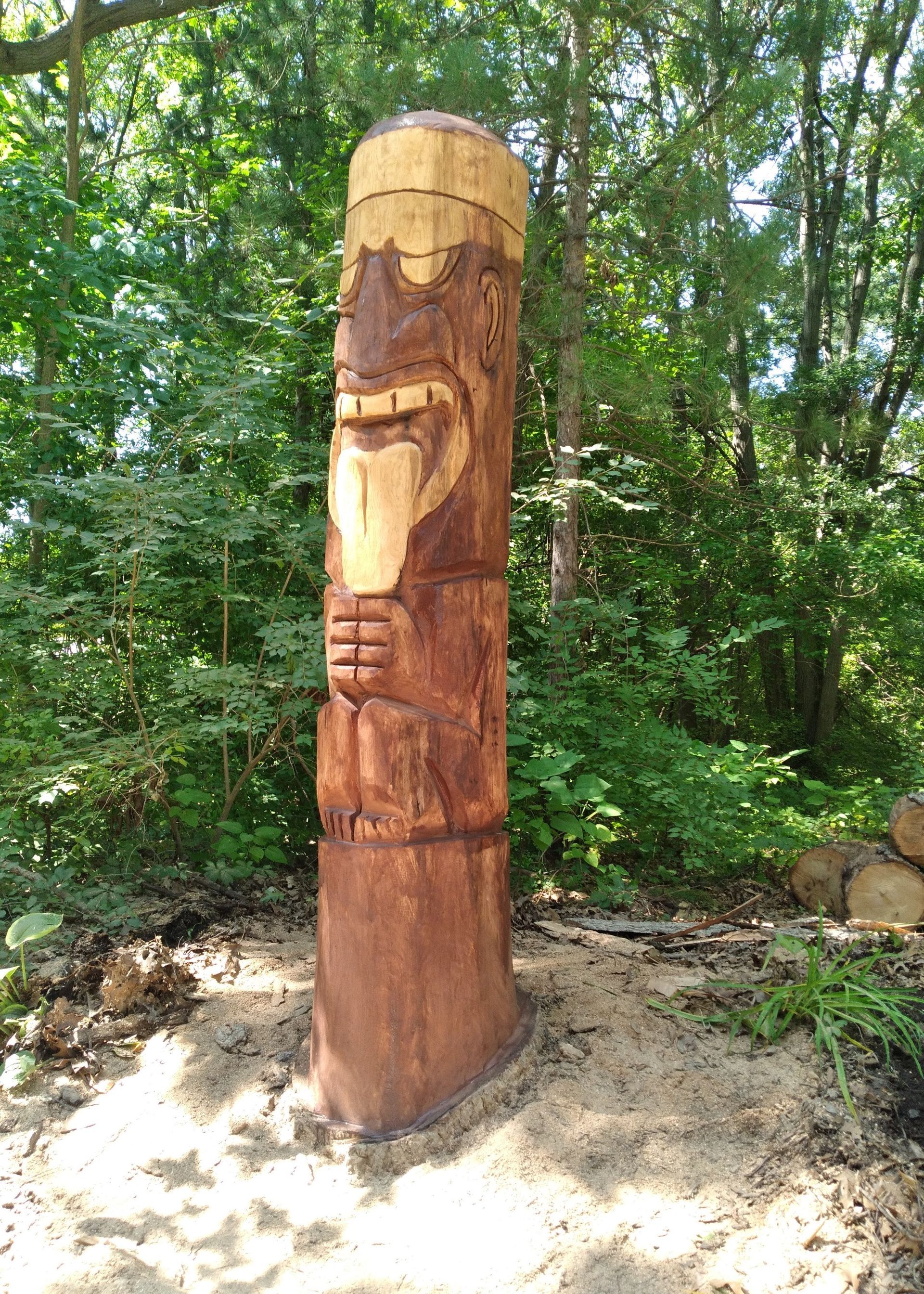 Wooden carved totem pole in a forest clearing, with green trees and sunlight in the background.