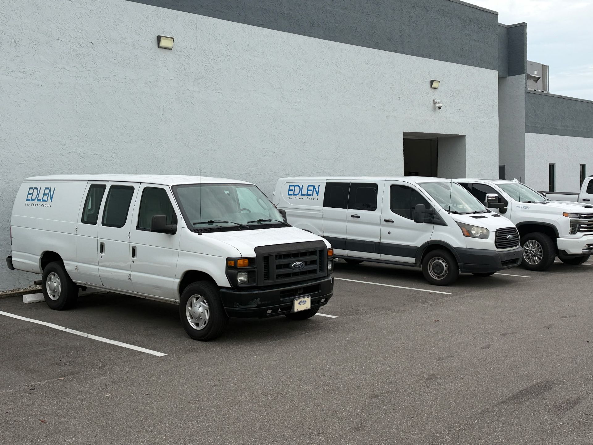 Row of white delivery vans parked diagonally on a paved lot Three white cargo vans parked outside a gray commercial building.