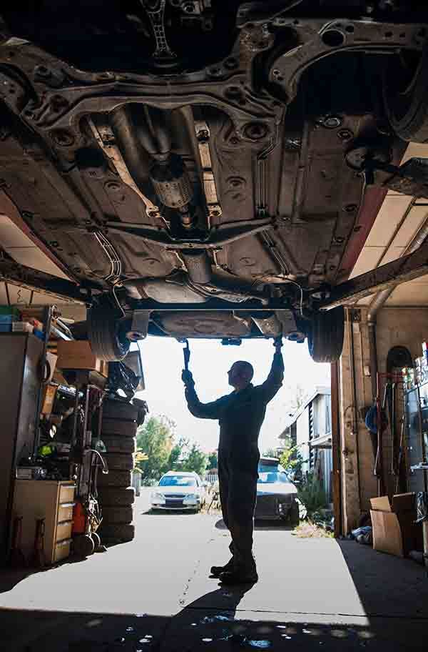 Mechanic silhouetted under a raised car in a garage, arms up inspecting the undercarriage.