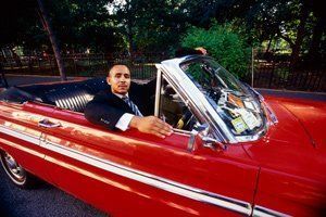 Man seated in a red convertible with white stripes, parked on a tree-lined street at dusk