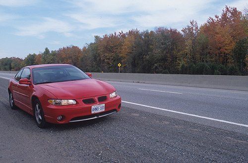 Red sports sedan parked on the shoulder of a highway beside autumn trees.
