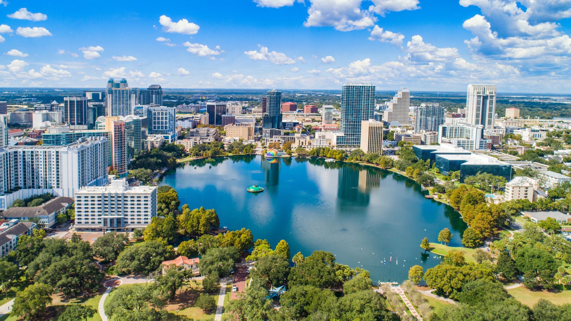 Mechanic working under a car lifted on a red hydraulic hoist in an auto repair shop Aerial view of a city skyline surrounding a blue lake, with parks and buildings under a bright sky