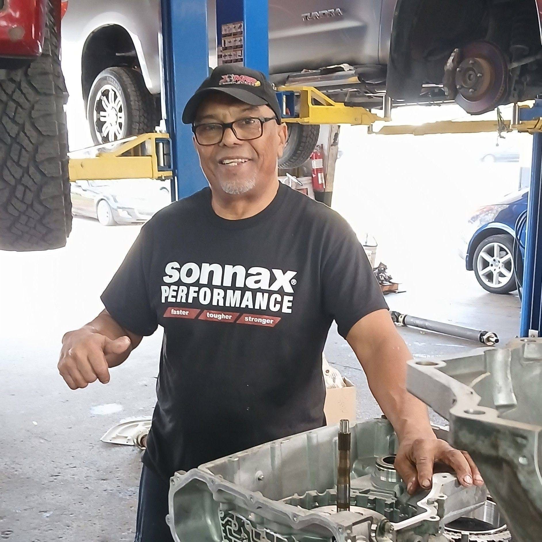 Mechanic in black Sonnax Performance shirt smiling in an auto shop beside a transmission on a workbench Mechanic in a Sonax Performance shirt standing in an auto shop beside an open engine block