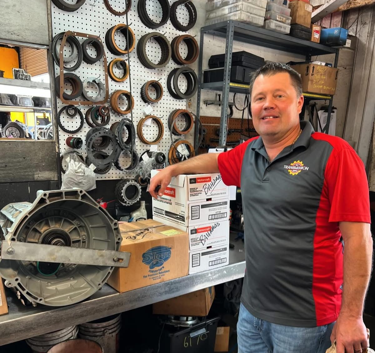 Man in red and gray shirt standing in auto parts shop beside boxes and a large metal engine part