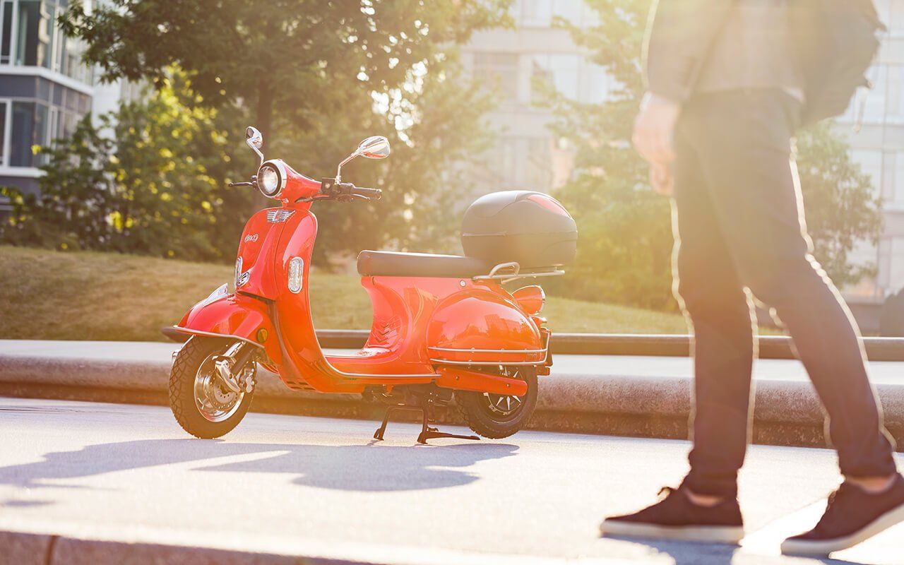 A person is walking past a red scooter on the side of the road.