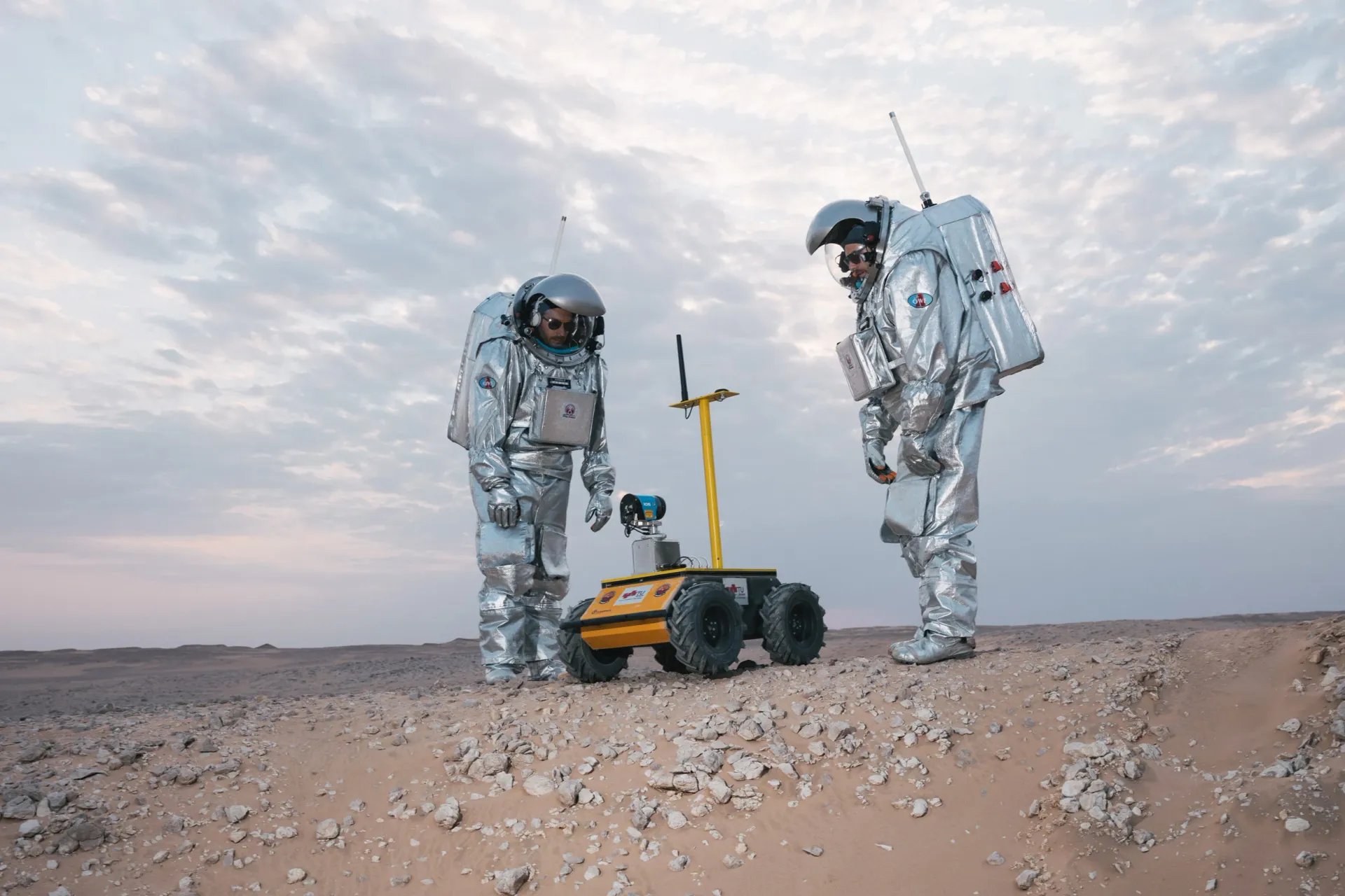 Two astronauts are standing next to a small rover in the desert.