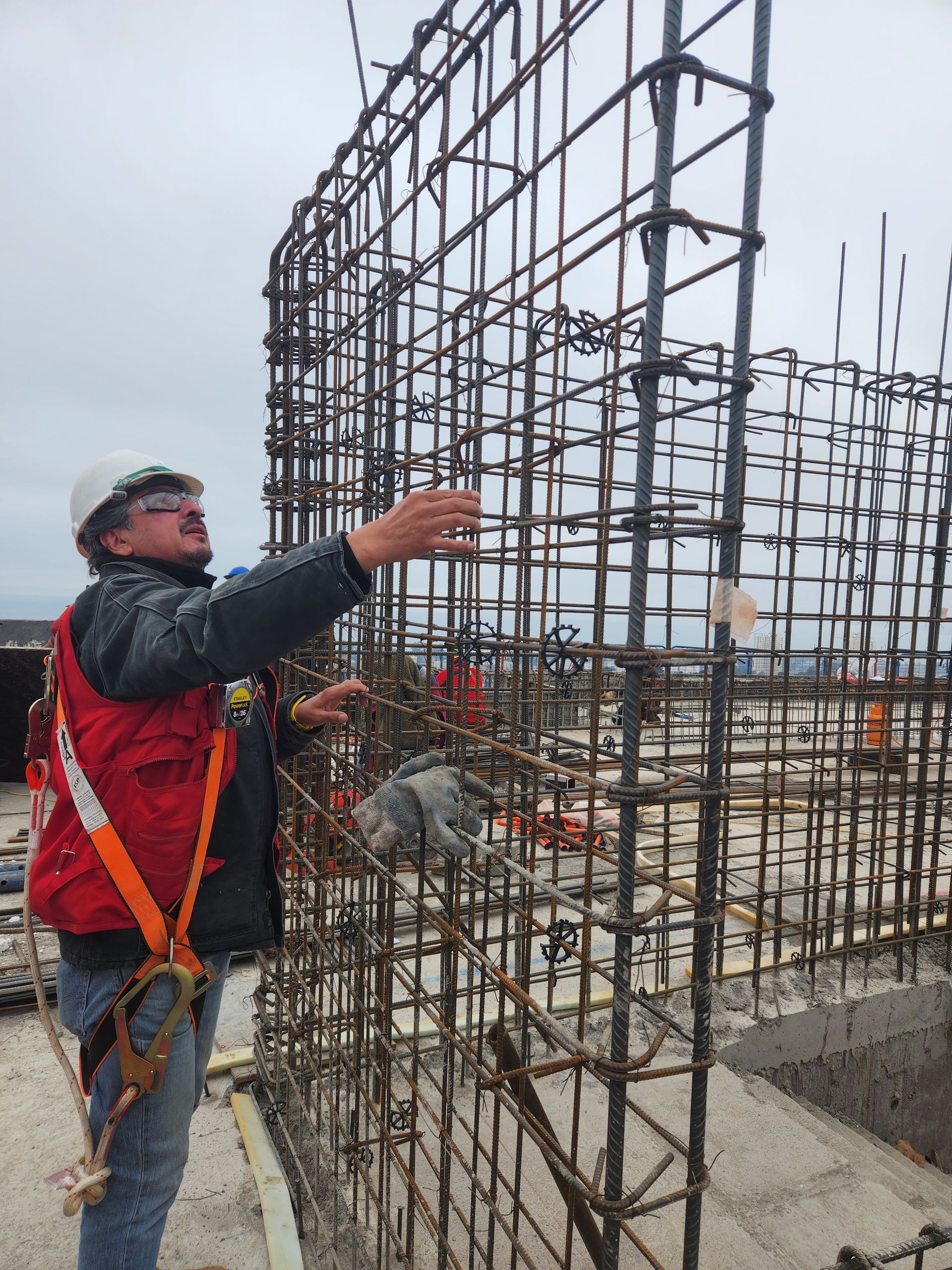 Obrero de la construcción inspeccionando una estructura de barras de acero en una obra bajo un cielo nublado.