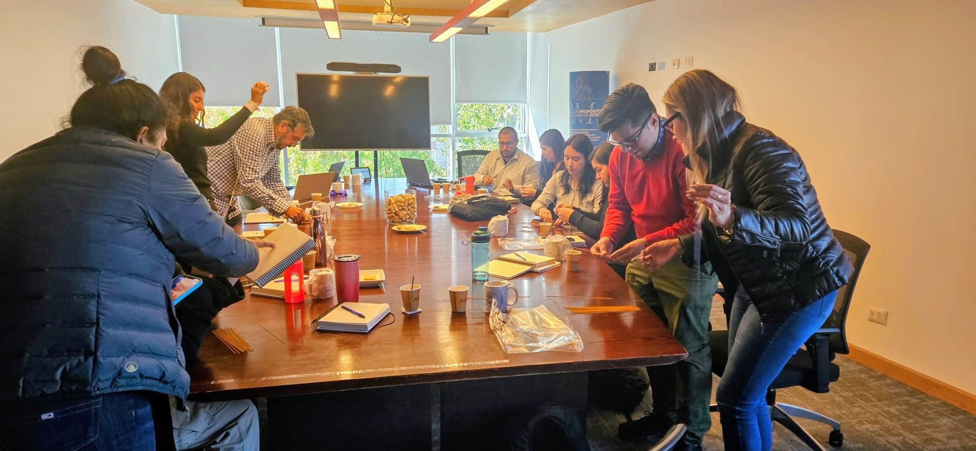 Varias personas se reunieron alrededor de una larga mesa en una habitación luminosa, comiendo y charlando durante una comida.
