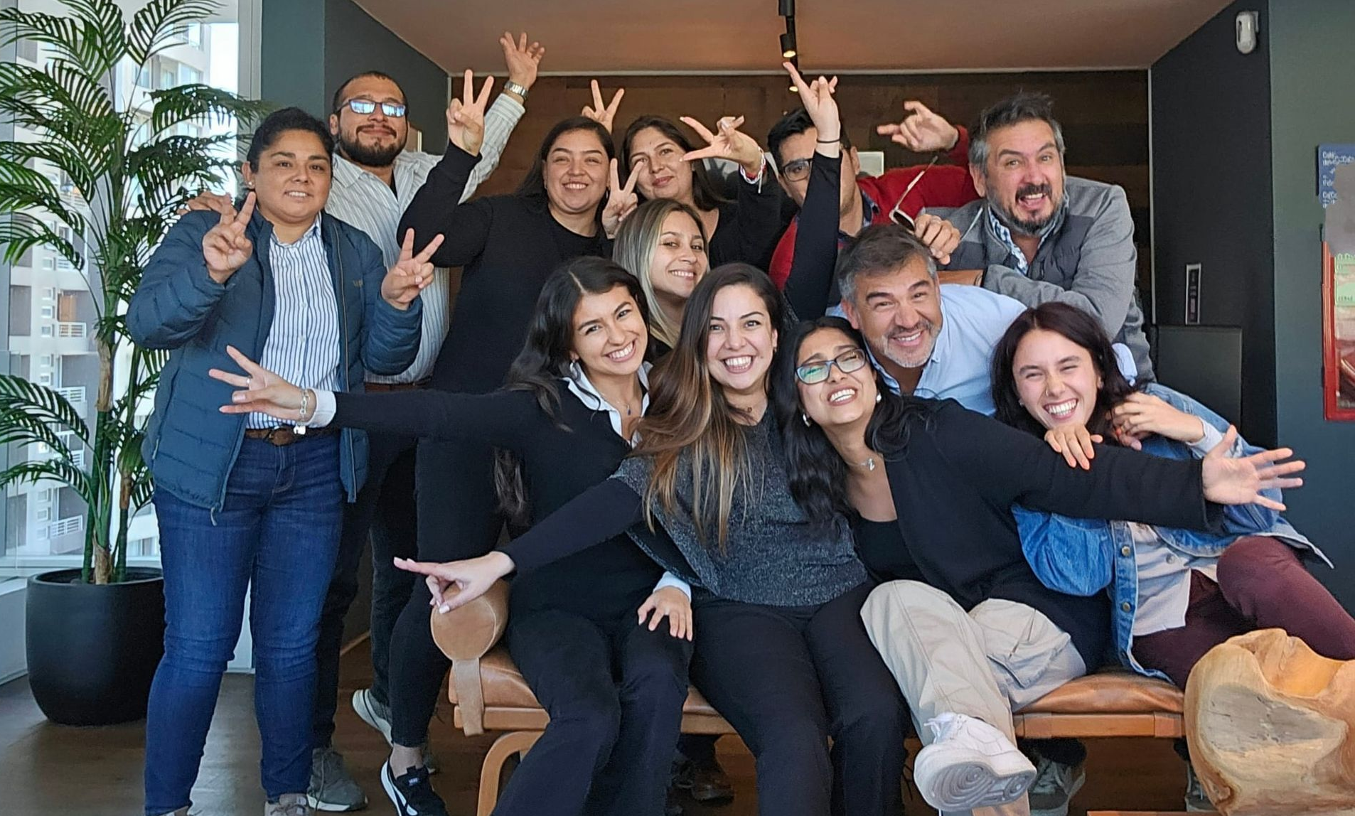 Grupo de personas posando y sonriendo en el interior, algunas sentadas y otras de pie, con las manos levantadas en un ambiente de oficina informal.