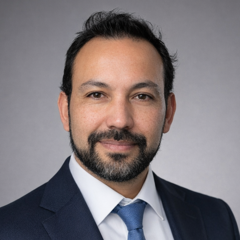 Retrato profesional de un hombre con traje oscuro, camisa blanca y corbata azul, sonriendo sobre un fondo gris.