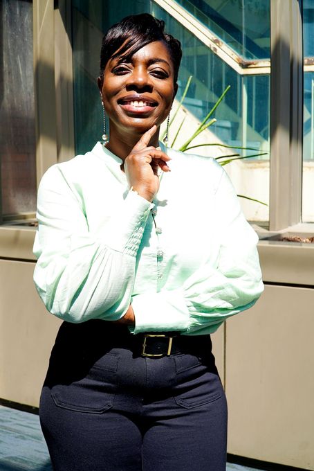 Person standing indoors by a window, smiling with arms crossed, wearing a light green blouse and dark pants.