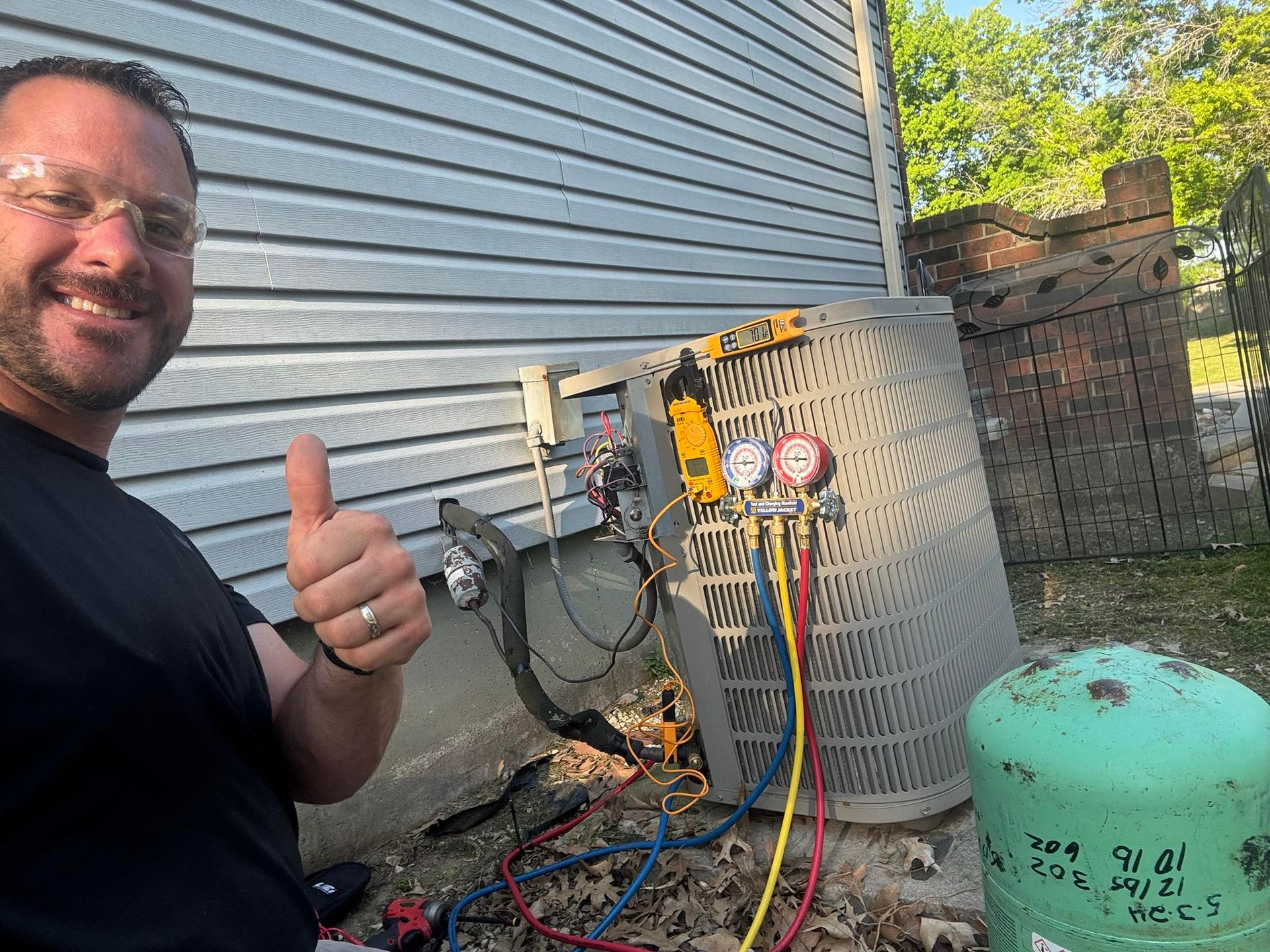 A smiling technician gives a thumbs up beside an outdoor air conditioning unit connected to pressure gauges.