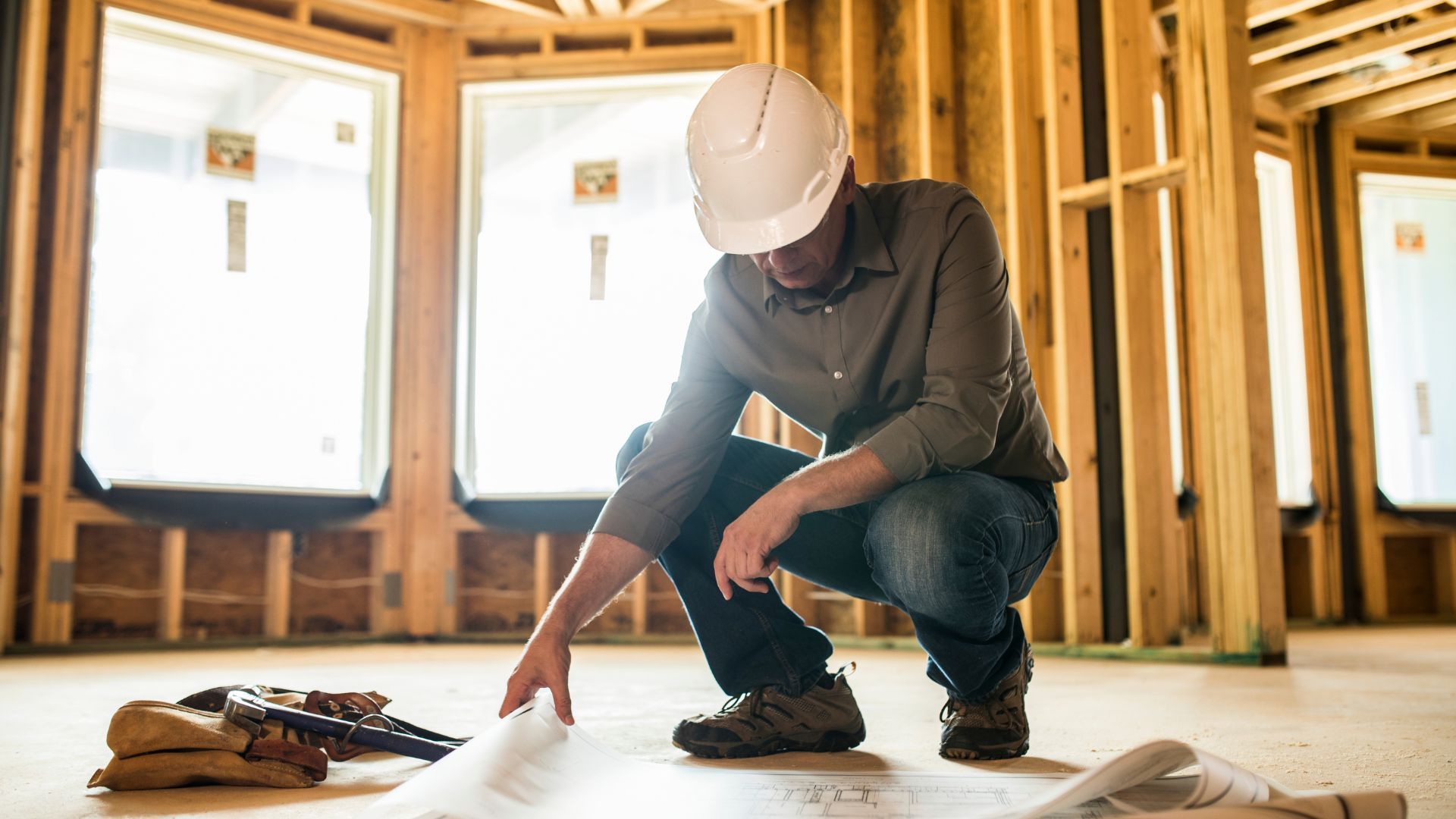 A construction worker is kneeling down in a room looking at a blueprint.