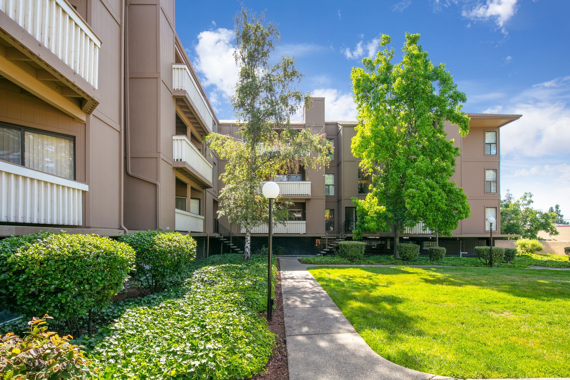 Apartment complex courtyard with green lawn, trees, shrubs, and a walkway under a bright blue sky at Vera West Valley in San Jose, CA.
