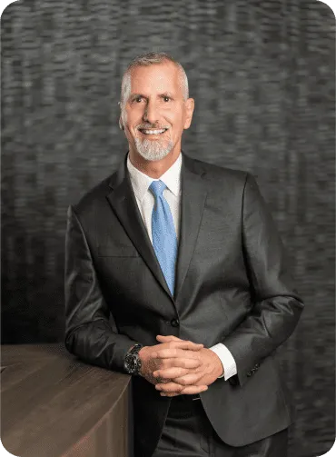 Man in a suit leans on a dark surface, smiling, with hands clasped; a blue tie and gray hair.