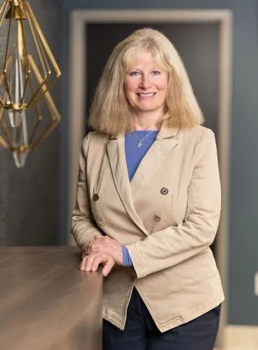 Woman in tan jacket smiles, leaning on a wooden counter with a decorative light fixture.
