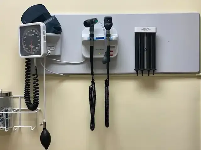 Medical exam equipment on a wall, including blood pressure monitor, otoscope, ophthalmoscope, and specula.
