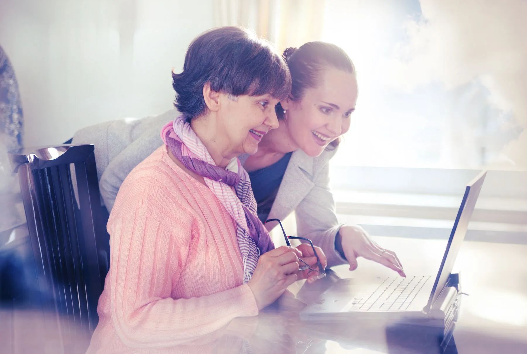 Woman in pink sweater and another woman looking at a laptop, both smiling. Bright, indoor setting.