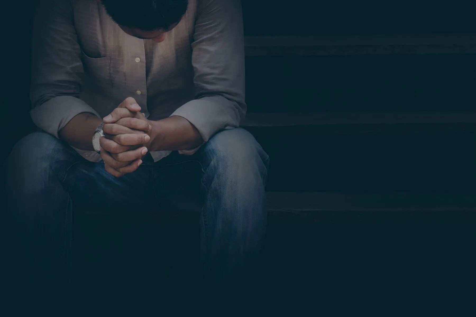 Man sits on steps, head bowed, hands clasped. Dark lighting creates a somber mood.