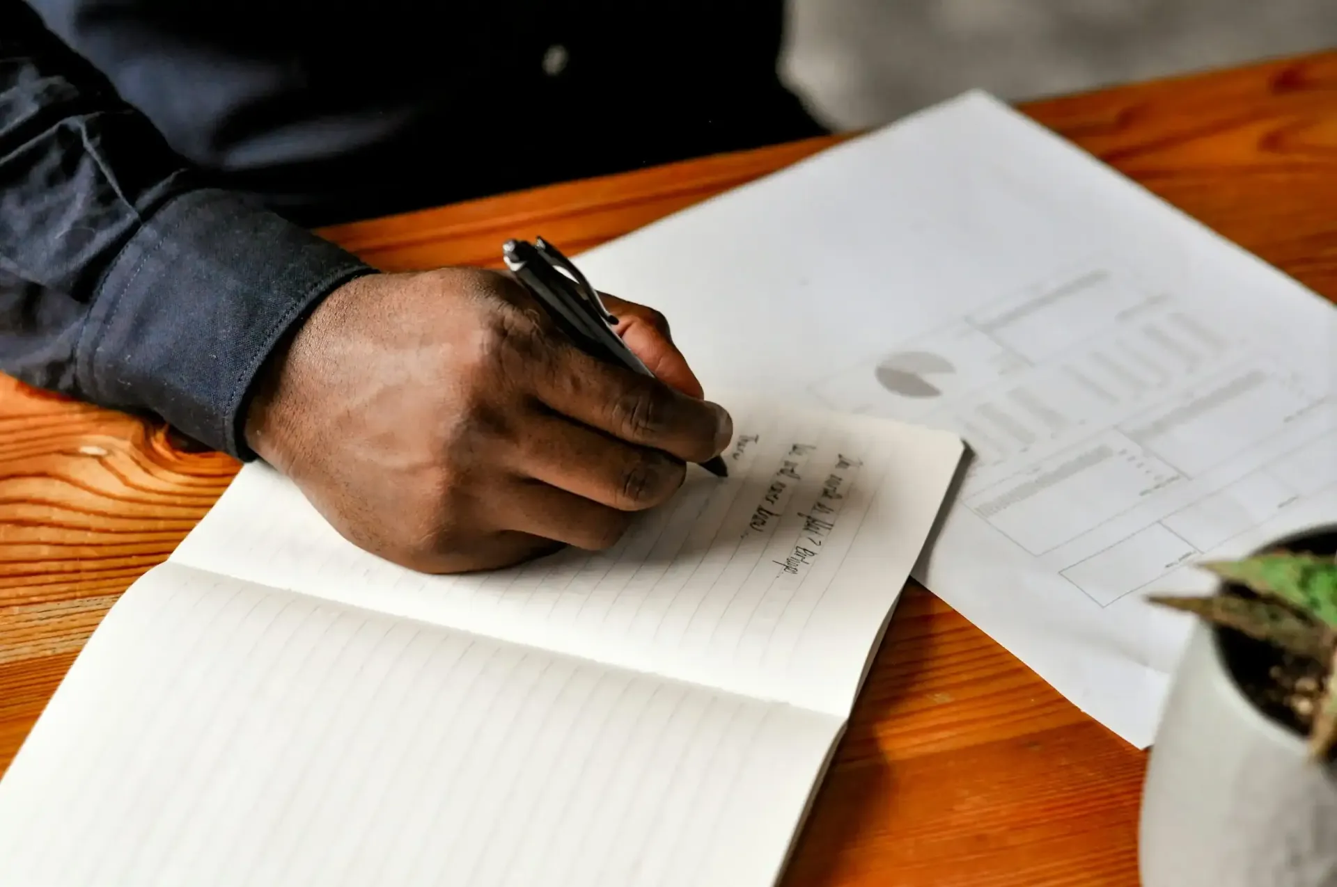 A person's hand writing in a notebook on a wooden table, another paper and plant are in the background.