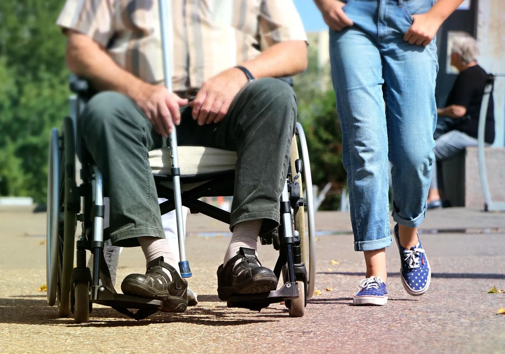 Man in wheelchair with person walking alongside on paved path.