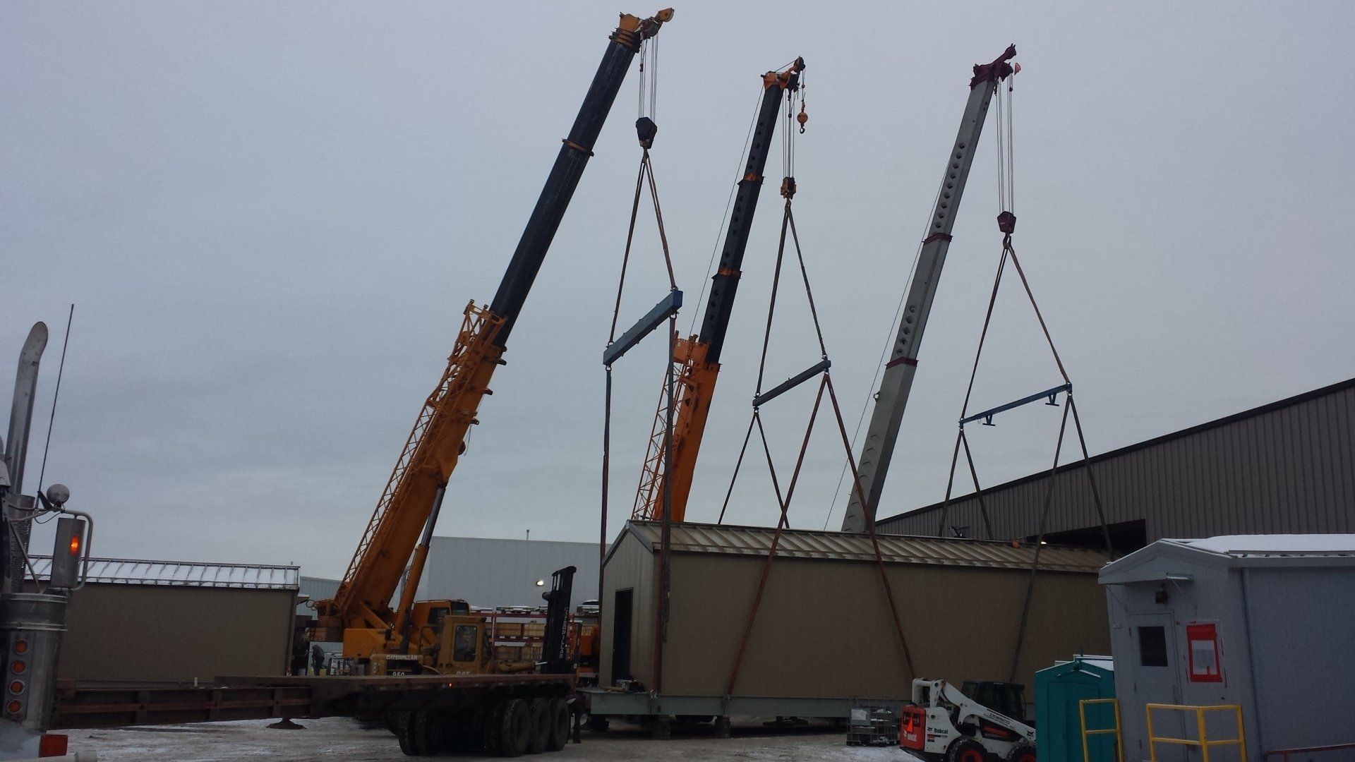 Three cranes lifting a modular building onto a flatbed truck on a snowy day.