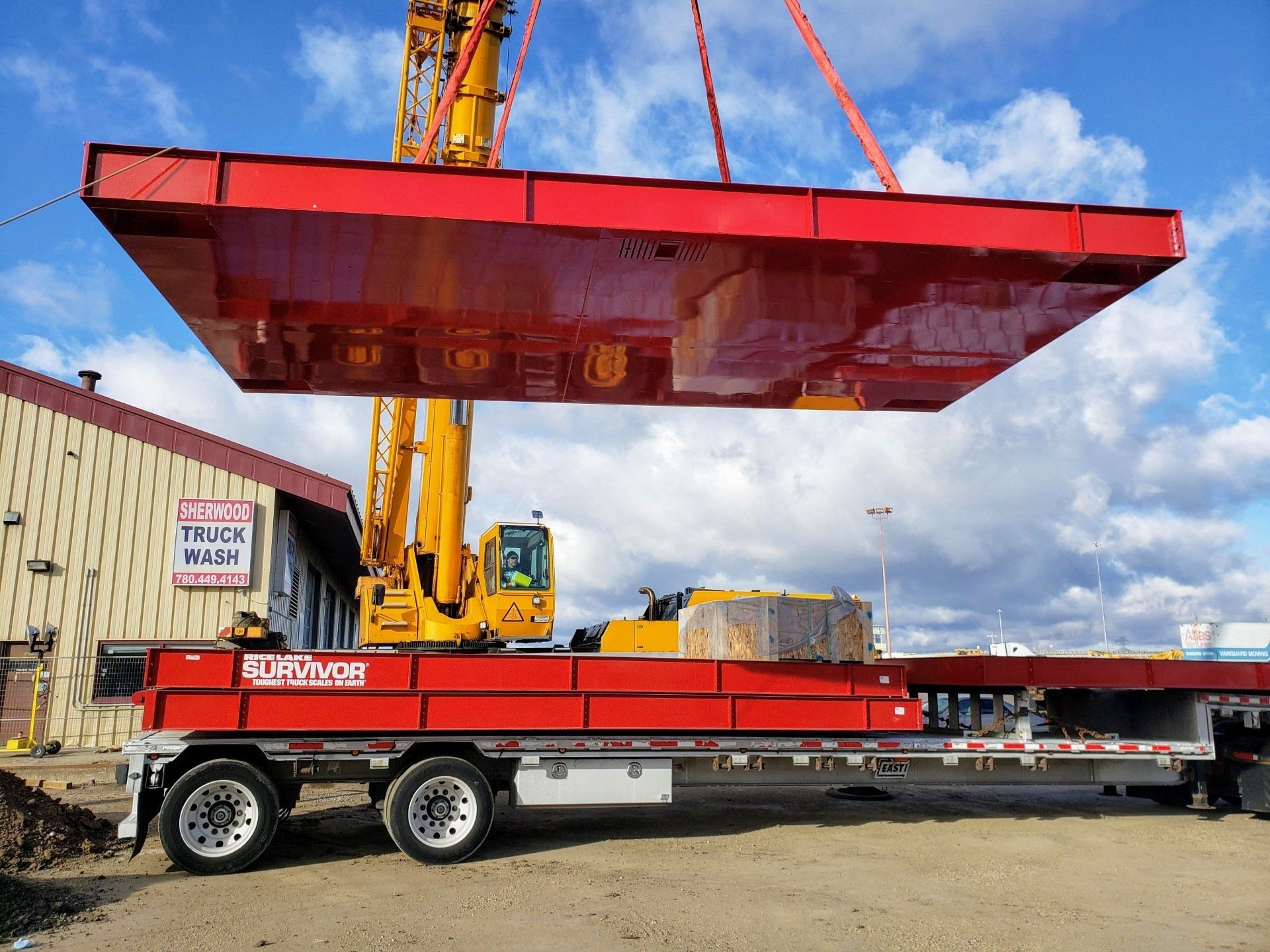 A large, red, rectangular structure being lifted by a crane over a trailer, outside a building.