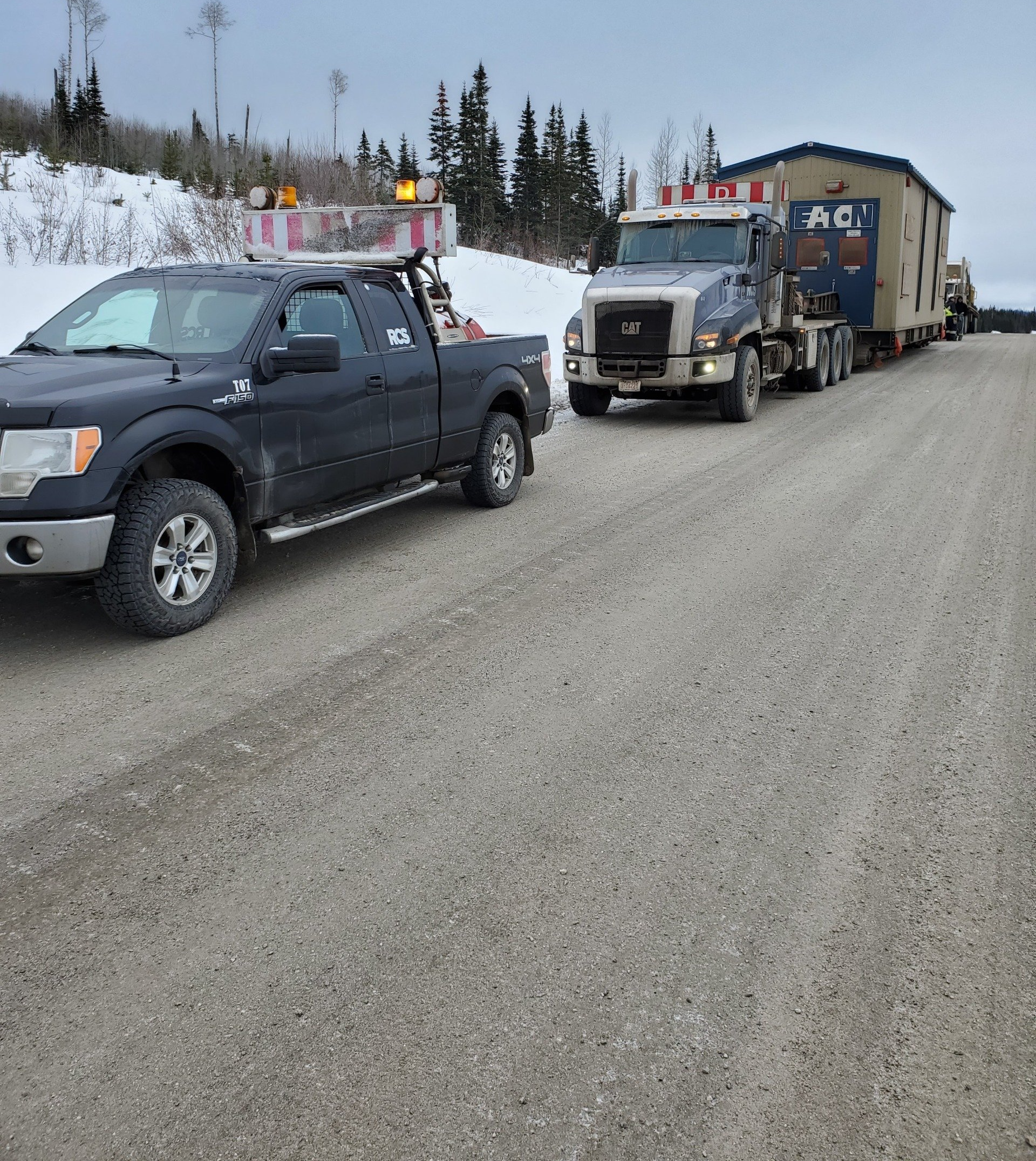 A black pickup truck and semi-truck hauling a modular building on a gravel road in a wintery setting.