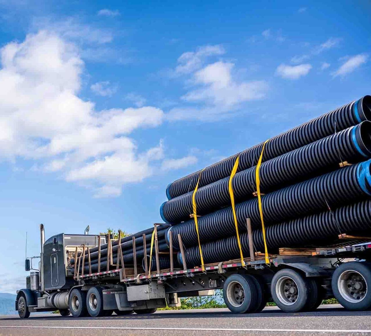 Semi-truck carrying a load of black corrugated pipes on a highway, under a blue sky.