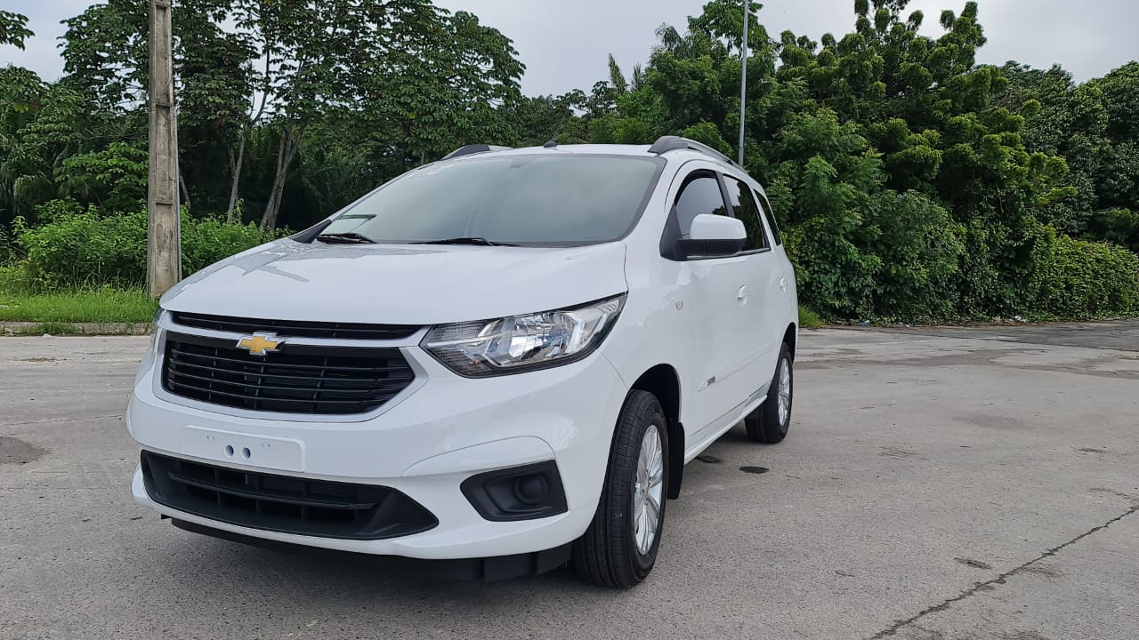 Um carro branco está estacionado em um estacionamento com árvores ao fundo.
