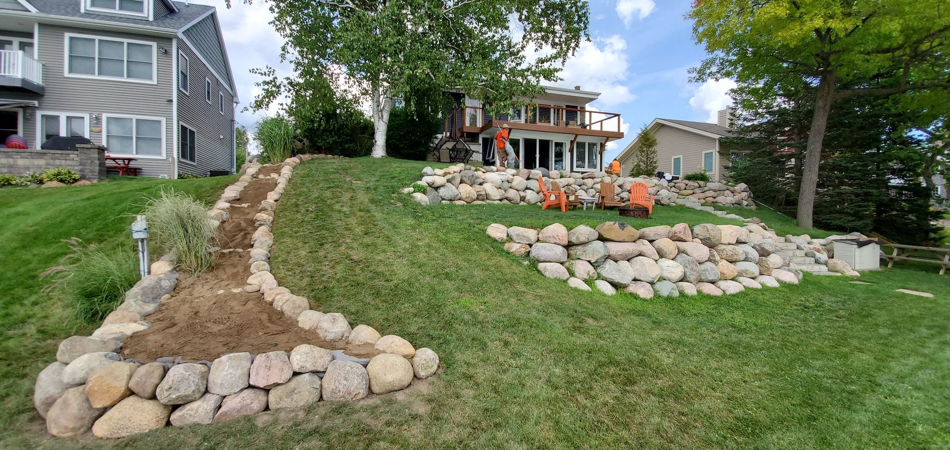 A grassy waterfront yard with stone retaining walls, a house, and a tree.