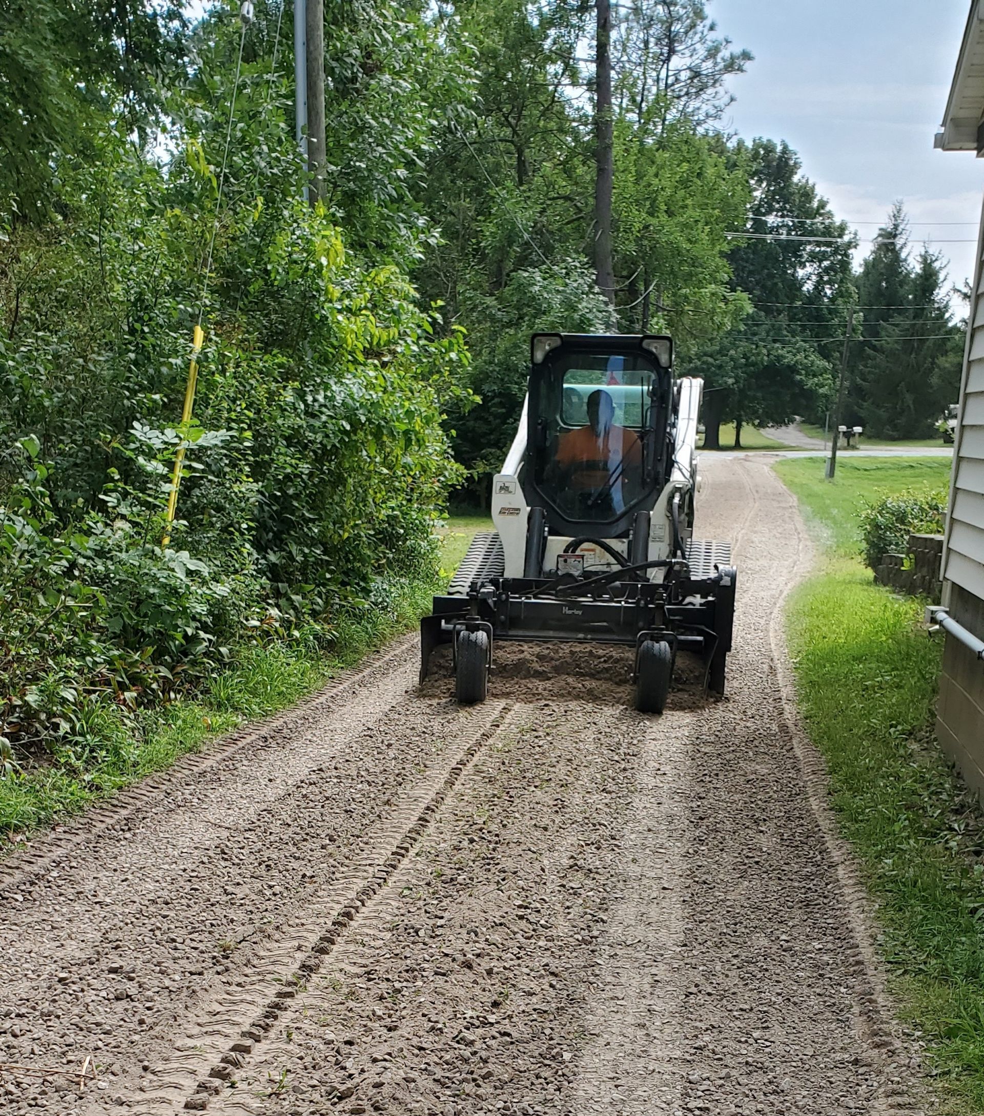 Bobcat leveling a gravel driveway, next to a house and trees.