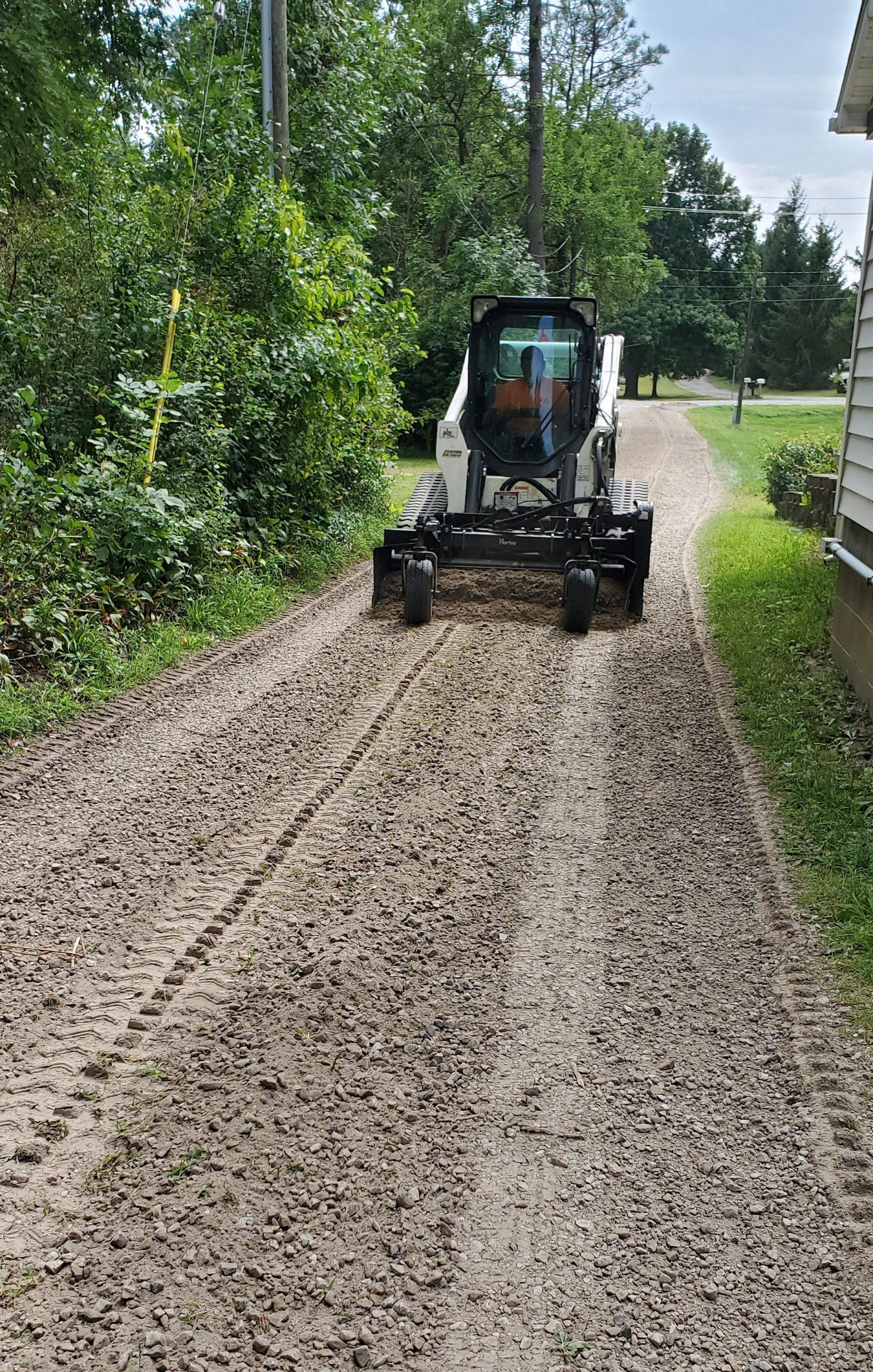 Bobcat skid steer operating on a muddy gravel driveway next to a house and trees.