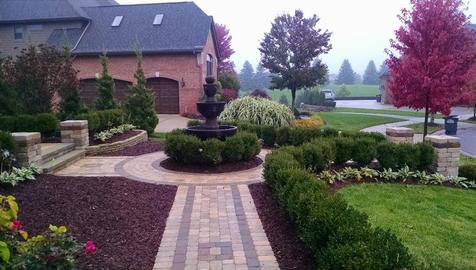 Brick pathway and landscaping lead to a water fountain in front of a large brick house with a cloudy, autumnal background.