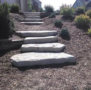 Stone steps ascending a sloped, mulched yard, surrounded by small bushes and greenery, leading to a house.