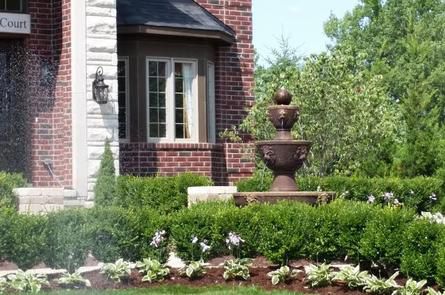 A fountain in a well-manicured garden in front of a brick house. Green hedges and white-edged plants surround the fountain.