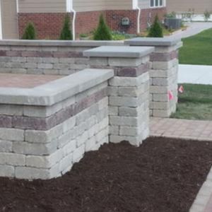 Brick retaining wall with decorative pillars and landscaping. Brown bricks alternate with beige bricks, topped with gray caps.